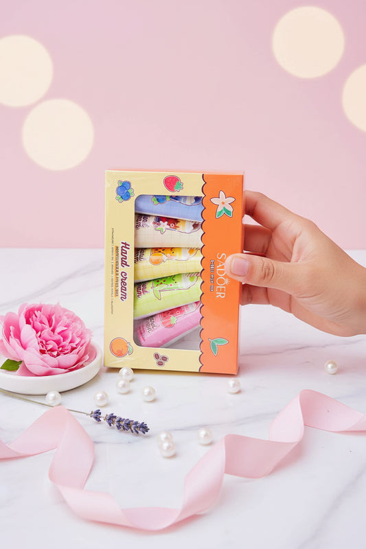 A hand holds a Sadoer hand cream set in its original packaging, featuring six tubes of hand cream visible through a clear window on a yellow and orange box, with a blurred green outdoor background.