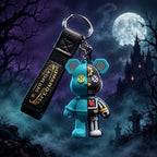 A hand holding a split-keychain charm featuring a half-blue bear and half-black-and-white robot, attached to a black strap with gold lettering, against a gray background.