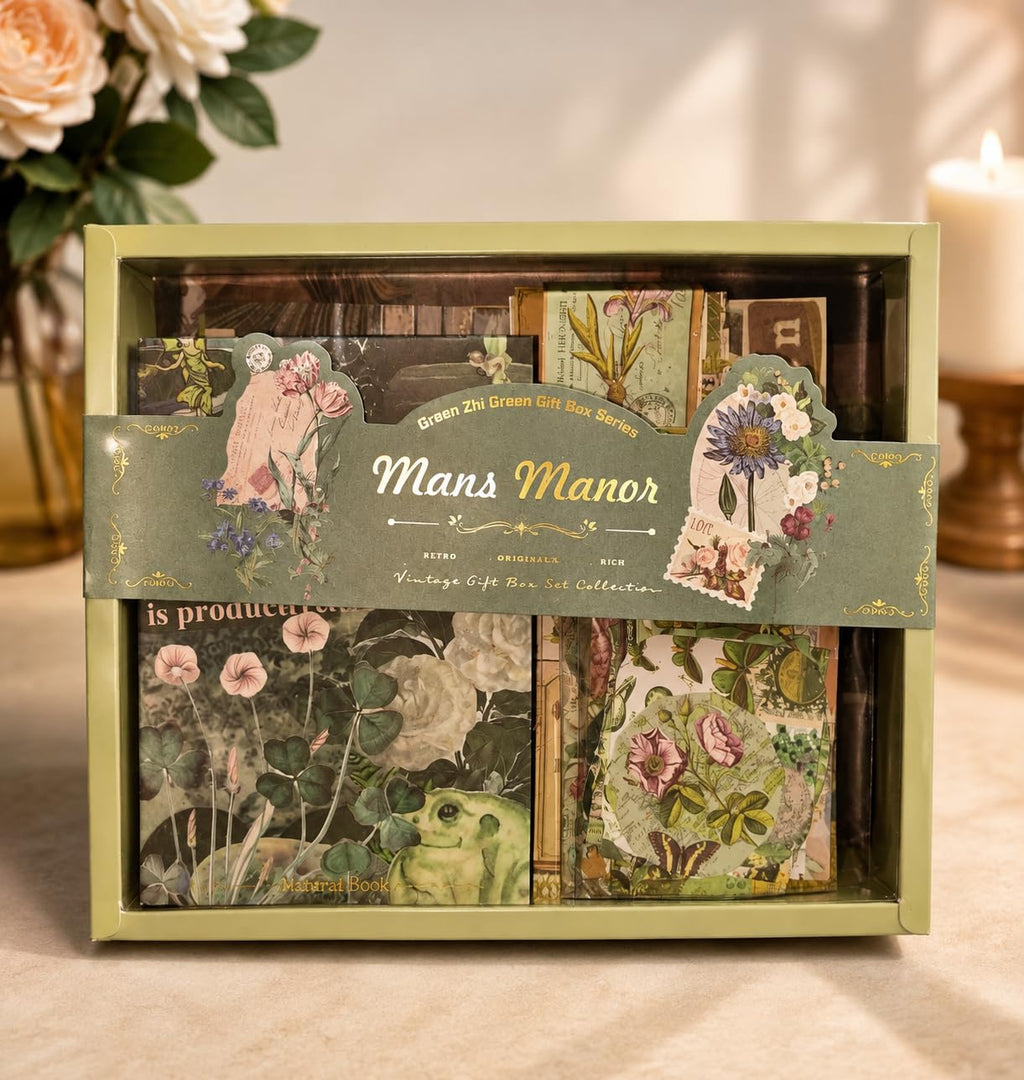 A boxed decorative card or scrapbook collection labeled "Mans Manor" with vintage-style floral illustrations, shown in a pale green box on a tabletop with flowers and a candle in the background.