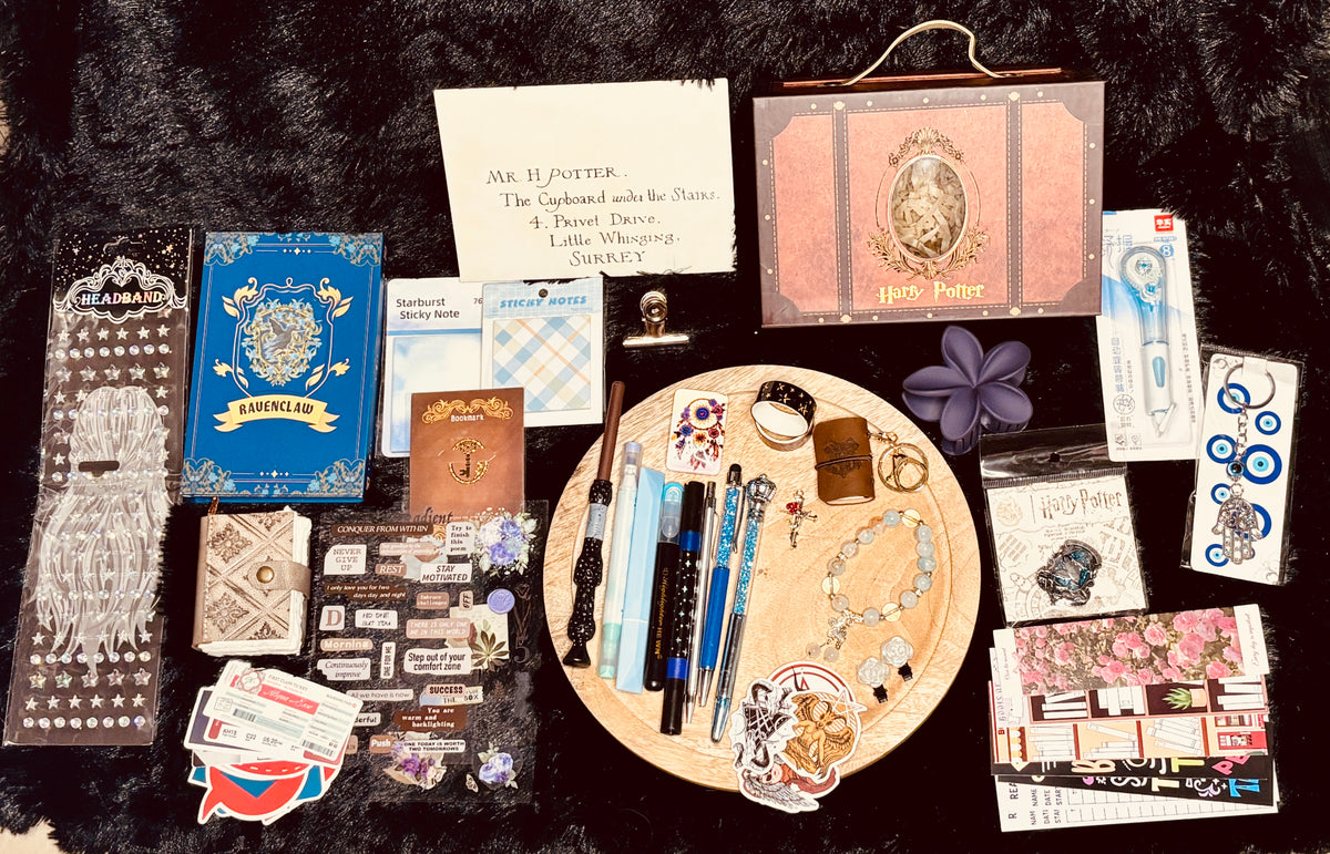 A styled flat-lay of Harry Potter themed stationery and accessories arranged on a black textured background. The collection includes a wooden tray with pens, stickers, and a bracelet, surrounded by books, cards, a notebook, and other themed items.