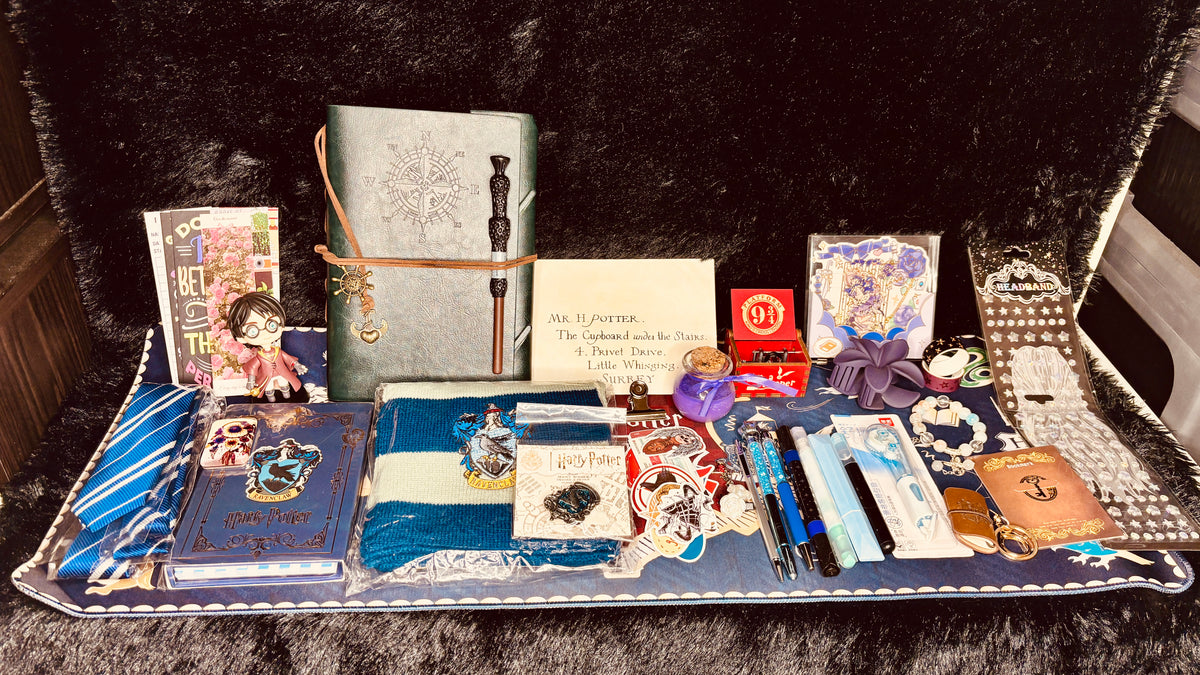 A curated collection of gothic-themed stationery and accessories arranged on a dark plush surface, including a gray journal with a wand, a blue notebook, a small doll, cards, pens, stickers, and other decorative items.