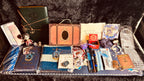A collection of Harry Potter themed merchandise arranged on a dark plush surface, including notebooks, a box, cards, pens, stickers, a small figurine, and other accessories.