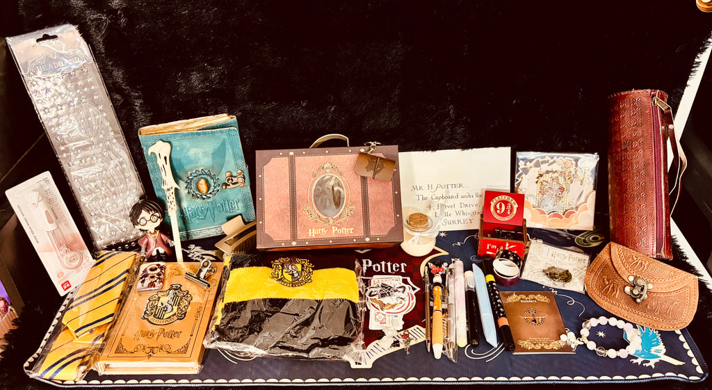 A curated collection of Harry Potter themed merchandise arranged inside a black suitcase against a dark background. The collection includes a replica Harry Potter trunk, a blue Hogwarts book, a yellow and black striped tie, a black robe, a brown leather pouch, a small red box, a white owl figurine, and other themed accessories.