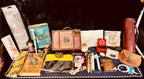 A curated collection of Harry Potter themed merchandise arranged inside a black suitcase against a dark background. The collection includes a replica Harry Potter trunk, a blue Hogwarts book, a yellow and black striped tie, a black robe, a brown leather pouch, a small red box, a white owl figurine, and other themed accessories.