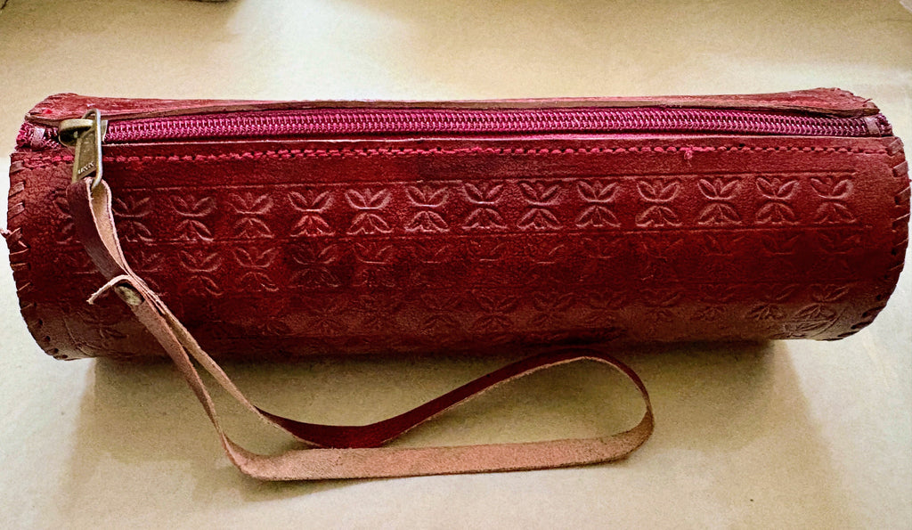 A cylindrical red leather zippered case with an embossed floral pattern and a light pink wrist strap, shown on a beige surface.