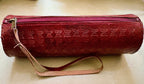 A cylindrical red leather zippered case with an embossed floral pattern and a light pink wrist strap, shown on a beige surface.