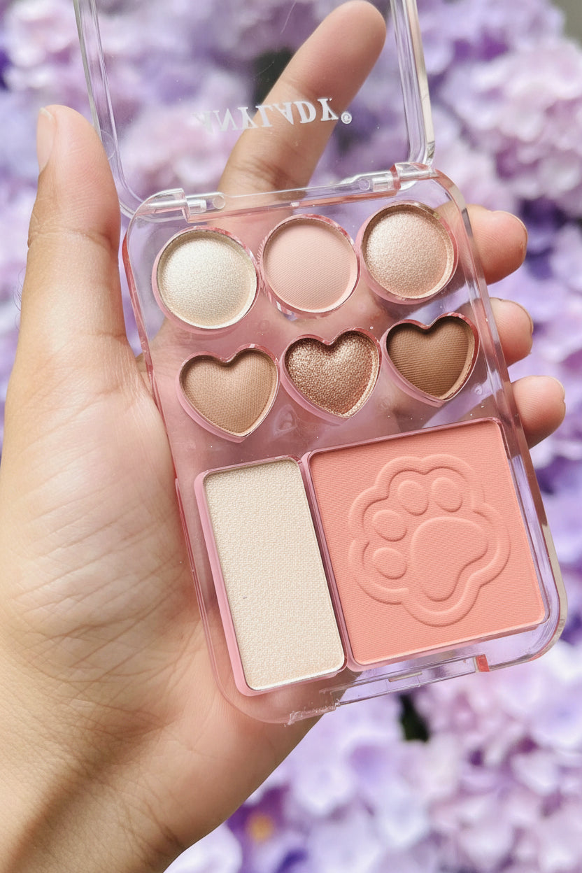 A hand holds a clear plastic makeup palette with six eyeshadow pans in round and heart shapes and a square blush compact with a paw-print embossed design. The background is a blurred green outdoor setting.