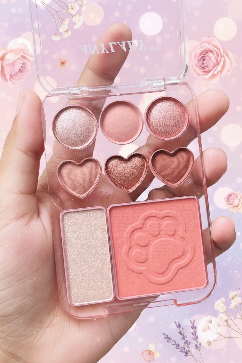 A hand holding a clear plastic makeup palette with six pink and rose-toned pans, including three round shimmer shades, two heart-shaped pressed powders, a rectangular shimmer pan, and a large pink paw-print shaped blush or highlighter. The background shows blurred green foliage.
