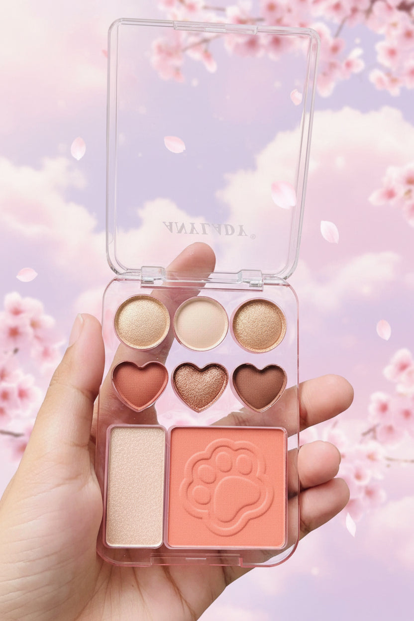 A hand holds a clear plastic makeup palette with six eyeshadow pans in warm neutral and peach shades, plus two square cream products, against a background of green outdoor foliage.
