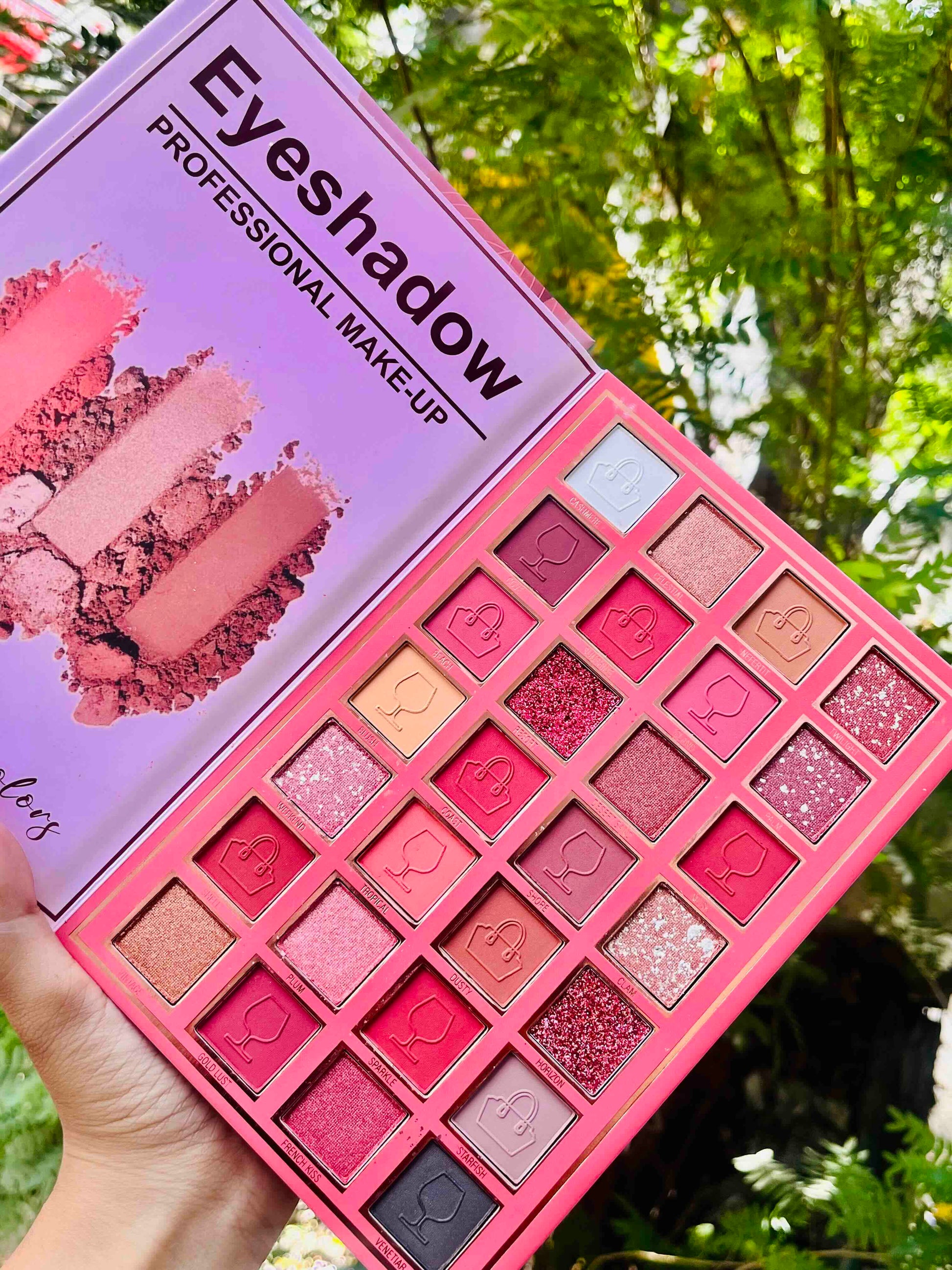 A pink eyeshadow palette held in a hand, showing 24 shades of eyeshadow in various matte, shimmer, and glitter finishes. The packaging features a lavender-purple background with text reading 'Eyeshadow PROFESSIONAL MAKE-UP' and swatches of makeup.