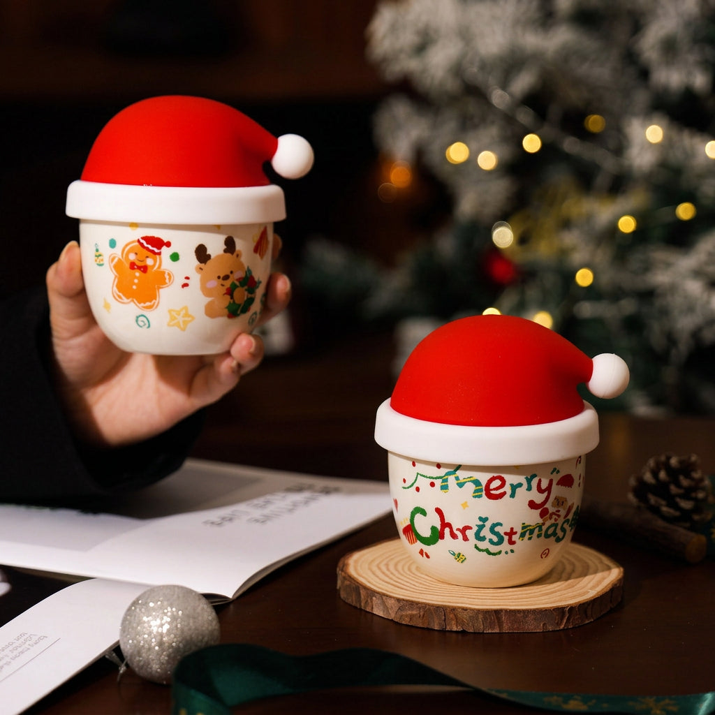 Two ceramic Christmas-themed cups with red Santa hats are shown on a wooden table in a cozy indoor setting with a blurred Christmas tree in the background. One cup is held by a hand, and the other sits on a wooden coaster.