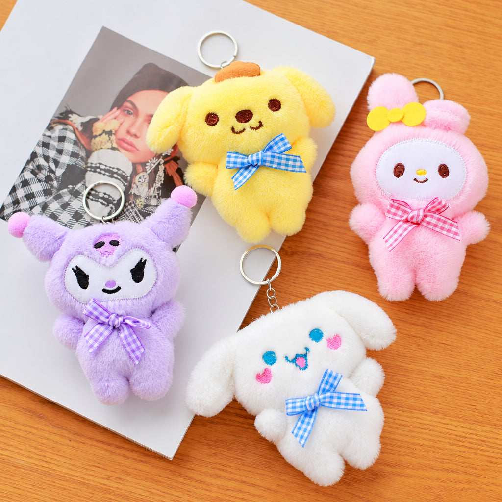 Four plush character keychains arranged on a wooden surface with a printed card underneath. The keychains are yellow, pink, purple, and white, each with a small bow and metal key ring.