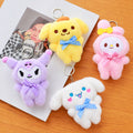 Four plush character keychains arranged on a wooden surface with a printed card underneath. The keychains are yellow, pink, purple, and white, each with a small bow and metal key ring.